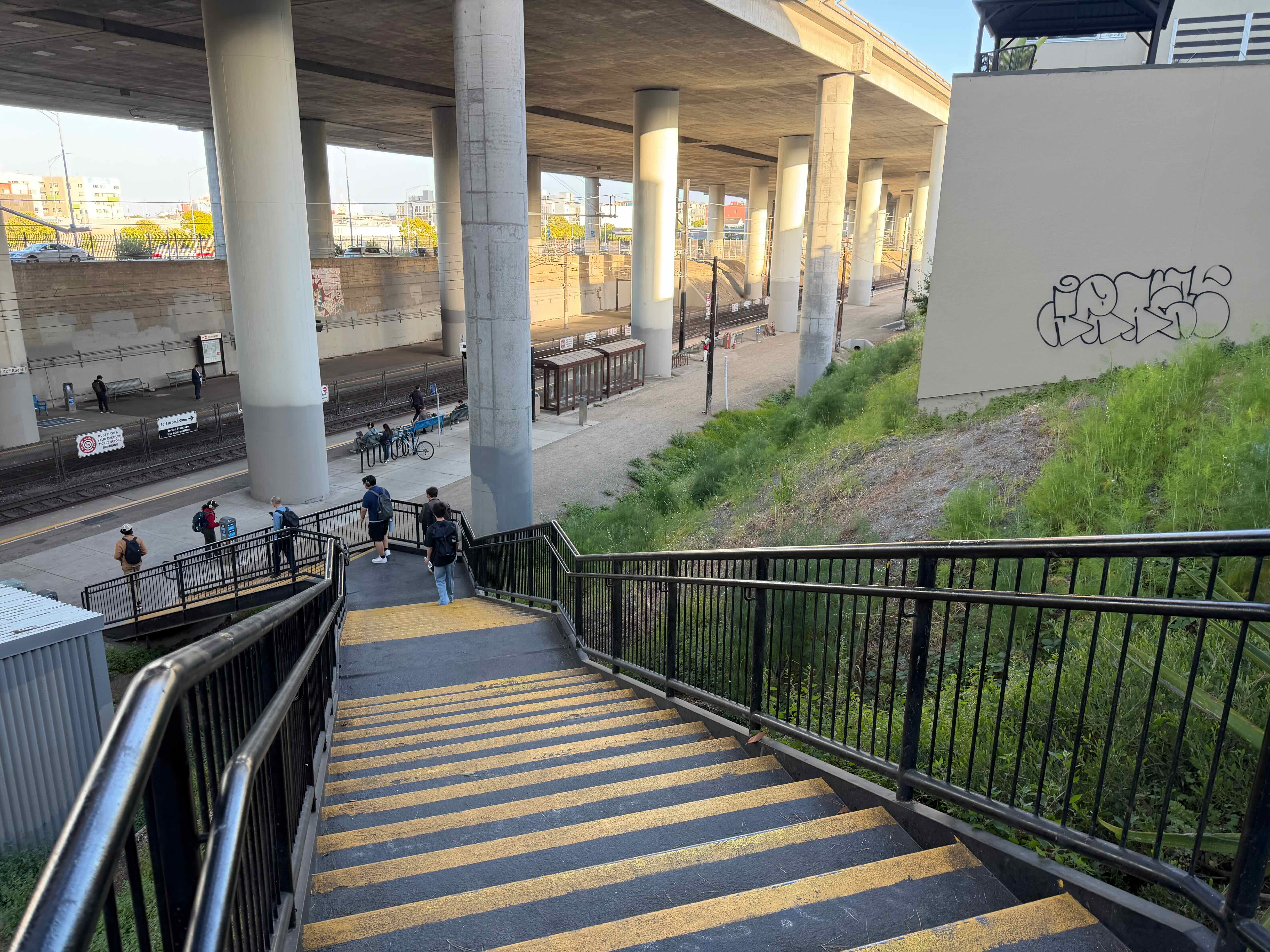 A view from the top of a tall staircase, separated into 3 flights. It descends into a large sunken area, at the bottom of which are the train tracks. A series of thick concrete pillars alongside the tracks hold up an elevated highway.