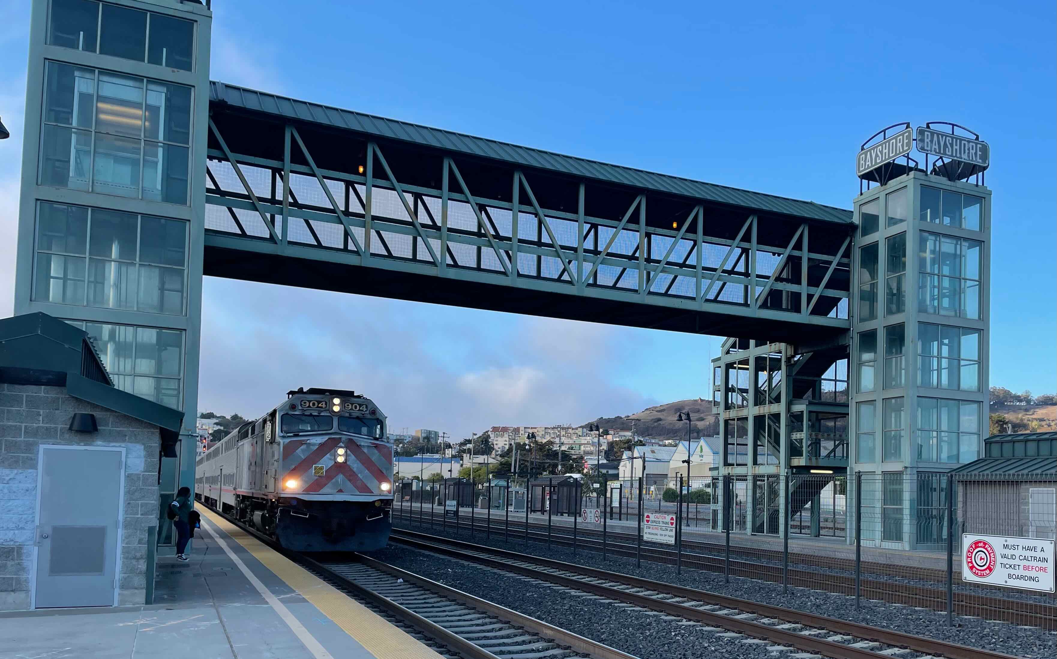 A train station with four tracks in the middle of two platforms. On each platform, a tower contains an elevator and three flights of stairs. A bridge spans the two towers over the tracks. A train is arriving at the station.