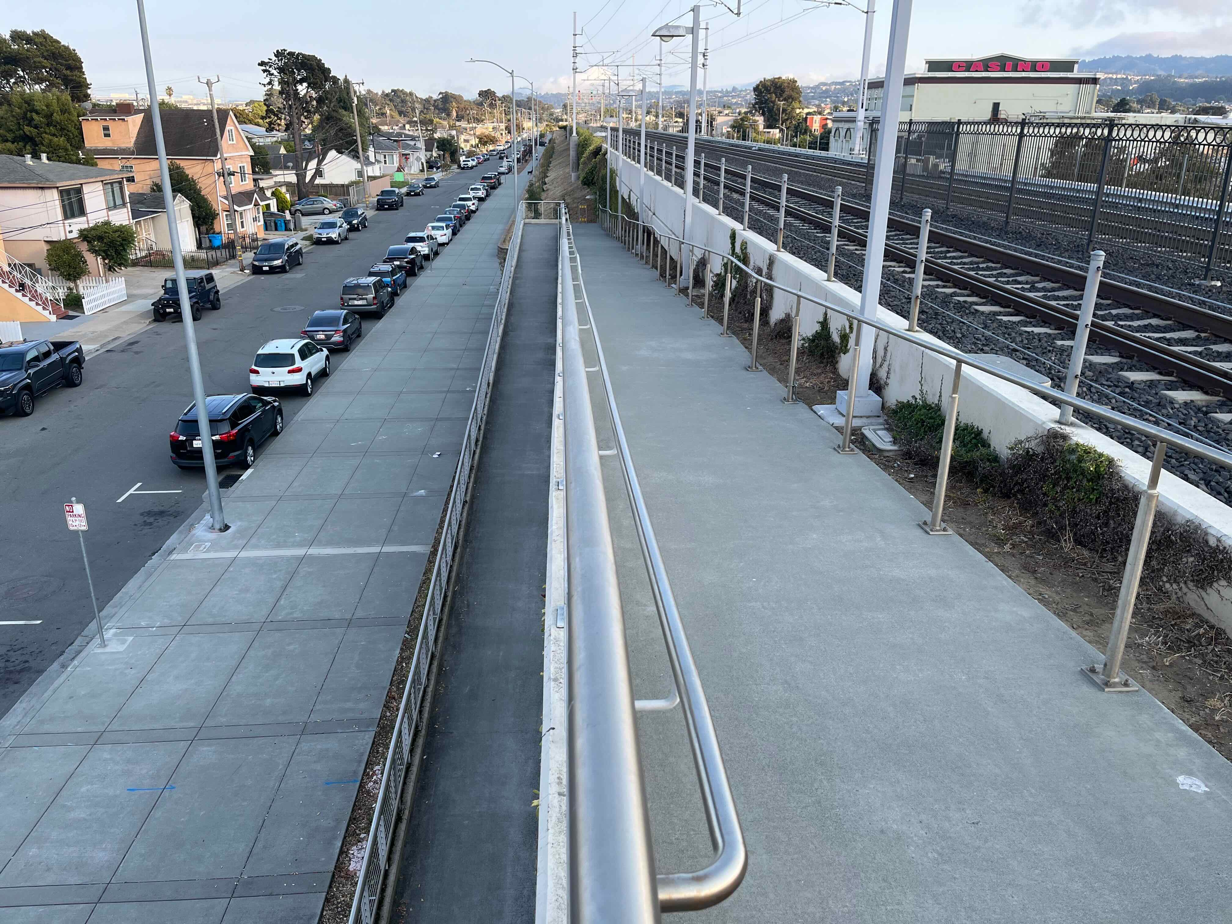 Along the side of a railroad embankment, a concrete ramp descends, with a 180degree switchback half of the way down. At the bottom of the embankment is a residential street with houses along the other side.