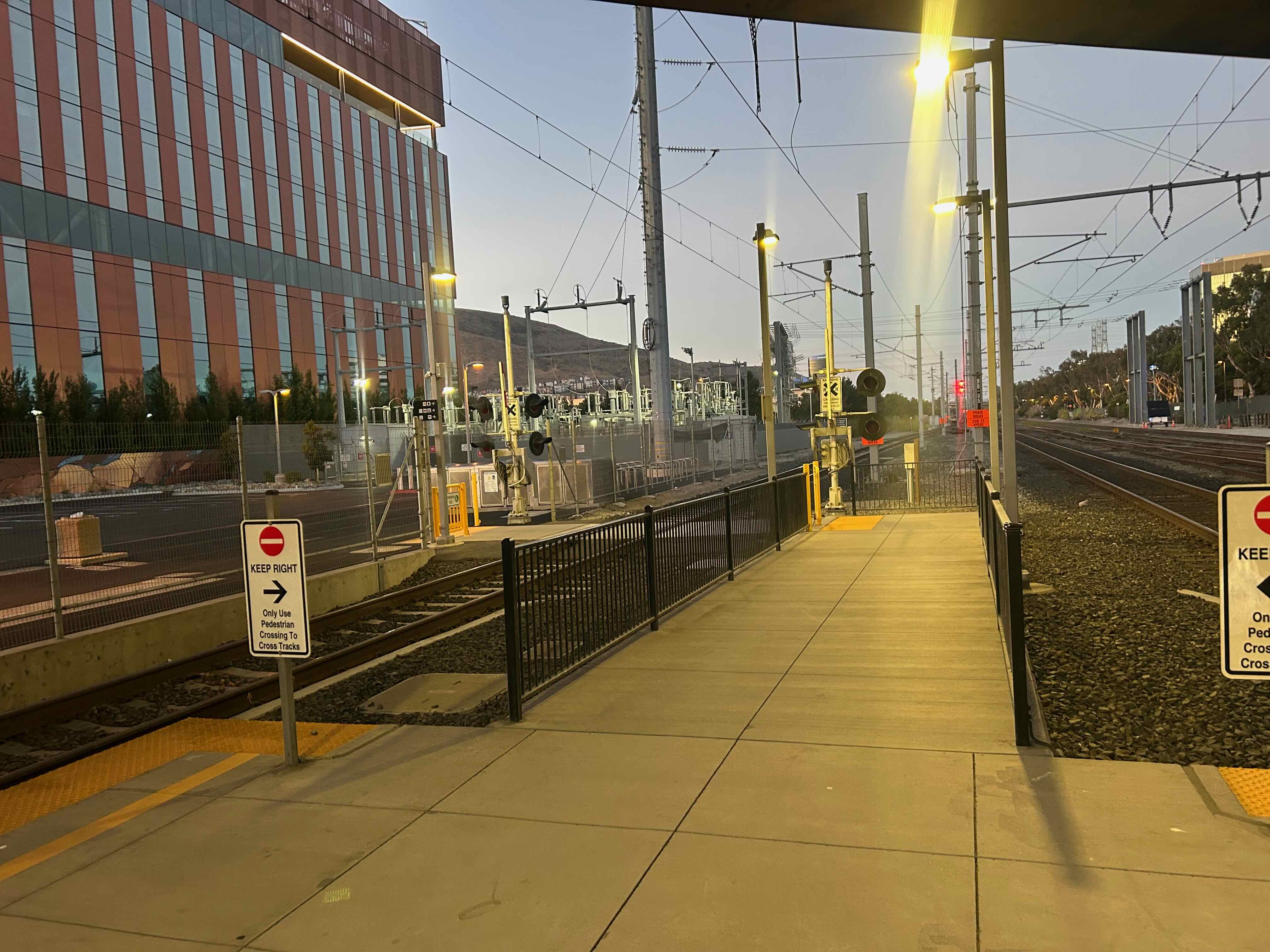 A concrete path extending about 5 meters from the end of a train platform between two tracks. The path has fences on both sides and at the end. At the end is an opening to the left, where the path crosses a track. The crossing has small crossing gates and lights, as well as yellow emergency exit gates. A parking lot is off to the left.