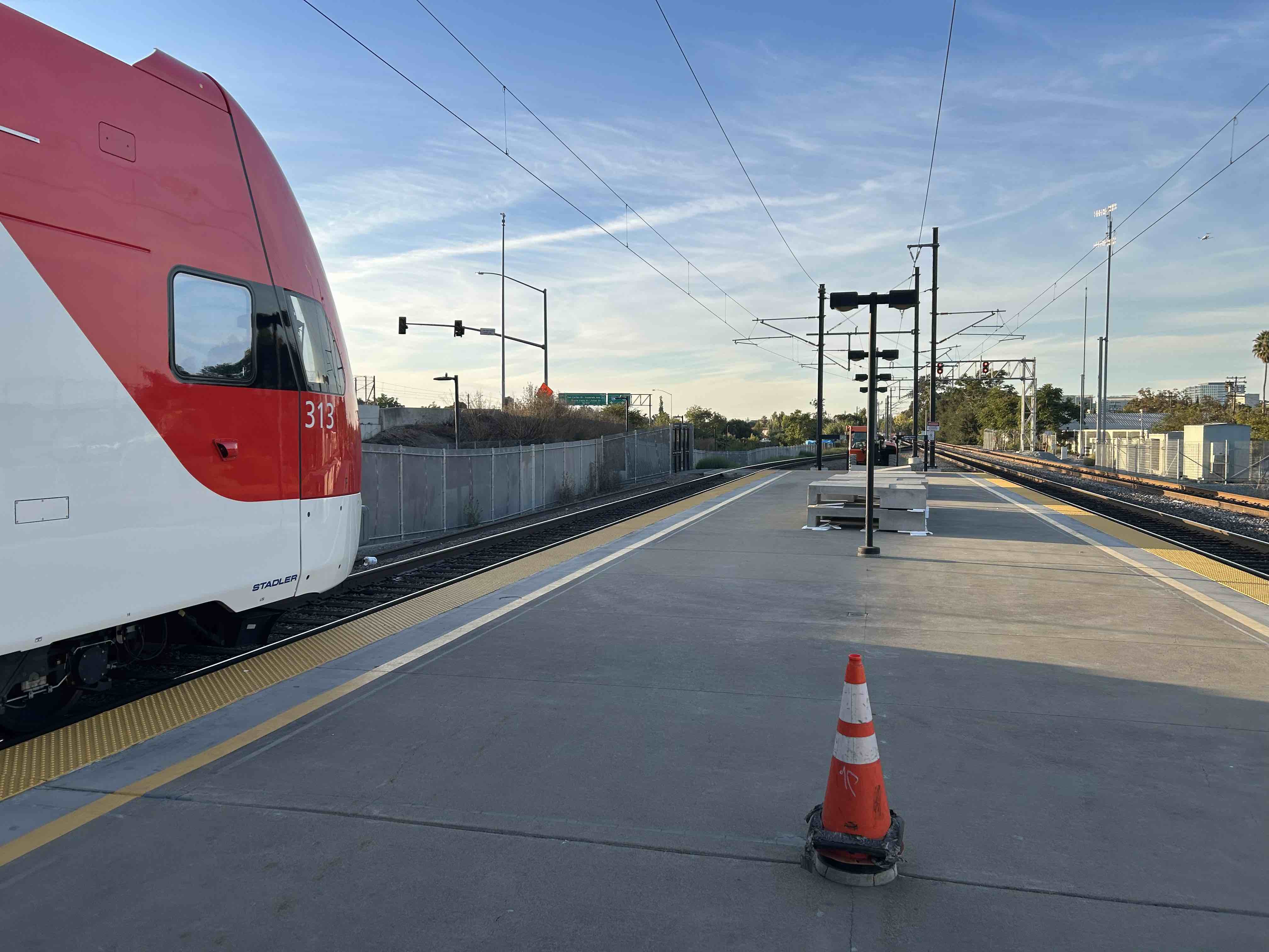 A view from near the end of a Caltrain platform, with two tracks running into the distance. Electrification wires are strung from poles between the tracks. The very front of a Caltrain EMU, #313, pokes out from the left edge of the photo. A construction cone is on the ground in the center of the platform.
